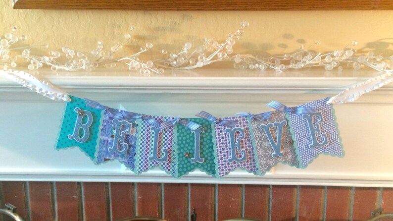 May include: A blue and white banner with the word "BELIEVE" spelled out in silver letters. The banner is made of paper and has a scalloped edge. It is hanging from a white string.