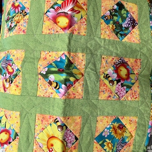May include: A green and yellow quilted fabric square with a diamond pattern. Each diamond features a different colorful floral design with bees, butterflies, and ladybugs.