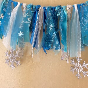 May include: A blue and white fabric banner with snowflake designs, ideal for a winter or Frozen themed party. The banner is made of various textures, including tulle, ribbon, and lace.