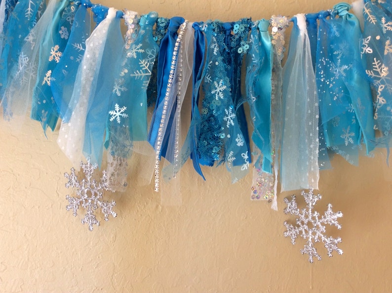 May include: A blue and white fabric banner with snowflake designs, ideal for a winter or Frozen themed party. The banner is made of various textures, including tulle, ribbon, and lace.