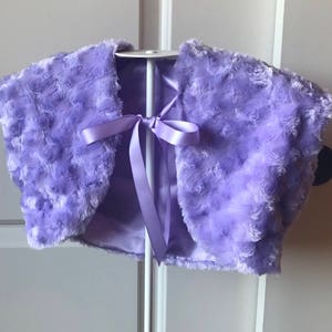 May include: A lavender faux fur shrug with a satin ribbon tie. The shrug is soft and fluffy, perfect for adding a touch of elegance to any outfit.