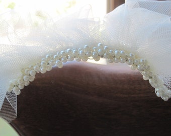 May include: Ivory-colored bridal headpiece featuring a delicate tulle veil and a curved band adorned with numerous small, round pearls. The veil is softly draped, creating a romantic and ethereal look.