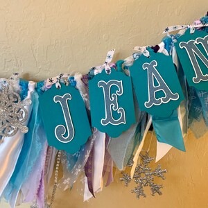 May include: A teal blue fabric banner with the name "JEAN" spelled out in white letters. The banner is decorated with white and blue ribbons, silver glitter snowflakes, and white lace.