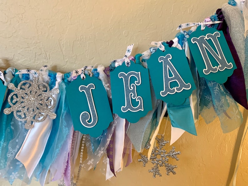 May include: A teal blue fabric banner with the name "JEAN" spelled out in white letters. The banner is decorated with white and blue ribbons, silver glitter snowflakes, and white lace.