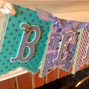 May include: A blue and green banner with white scalloped edges and the word "BELIEVE" in white letters. The banner is decorated with blue and white polka dots, snowflakes, and a blue and white gingham ribbon.