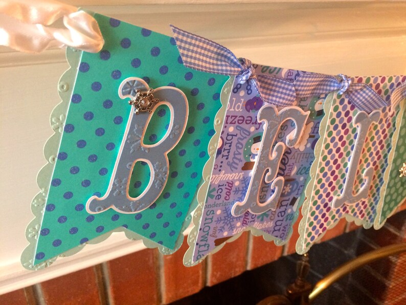 May include: A blue and green banner with white scalloped edges and the word "BELIEVE" in white letters. The banner is decorated with blue and white polka dots, snowflakes, and a blue and white gingham ribbon.