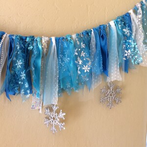 May include: A blue and white fabric garland with snowflake designs. The garland is made of various textures, including tulle, ribbon, and lace.