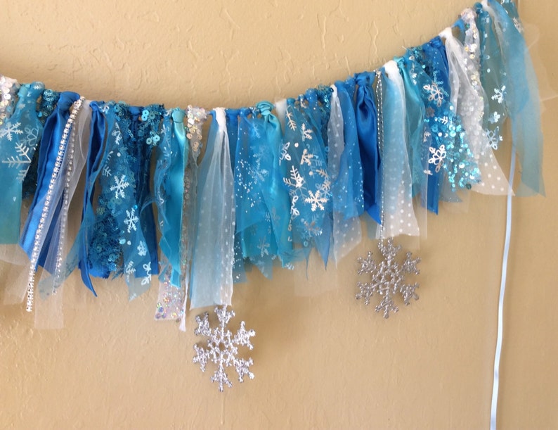May include: A blue and white fabric garland with snowflake designs. The garland is made of various textures, including tulle, ribbon, and lace.