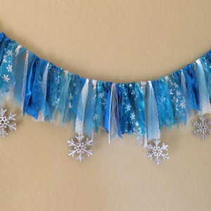 May include: A blue and white fabric banner with silver snowflake accents. The banner is made of various textures of fabric, including tulle, ribbon, and satin.