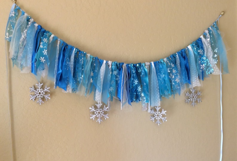 May include: A blue and white fabric banner with silver snowflake accents. The banner is made of various textures of fabric, including tulle, ribbon, and satin.