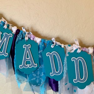 May include: A teal blue fabric banner with the name "MADDY" spelled out in white letters with a glittery finish. The banner is decorated with white and blue snowflake patterned fabric, purple satin ribbon, and white tulle.
