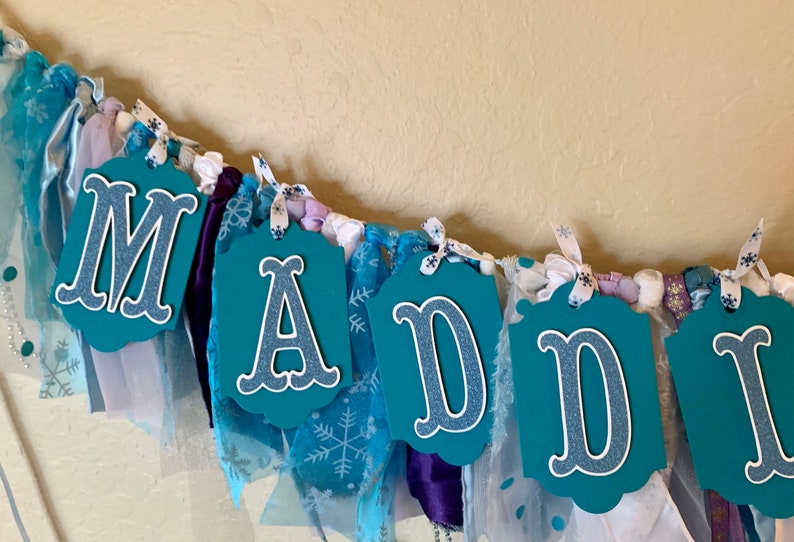 May include: A teal blue fabric banner with the name "MADDY" spelled out in white letters with a glittery finish. The banner is decorated with white and blue snowflake patterned fabric, purple satin ribbon, and white tulle.