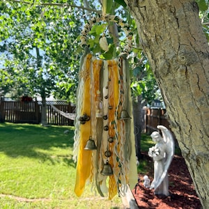 May include: A circular wall hanging featuring a wrapped hoop adorned with yellow and olive green fabric ribbons, yarn, and twine. The piece includes hanging metal bells, a small glass bottle containing sand, and a central crystal pendant.
