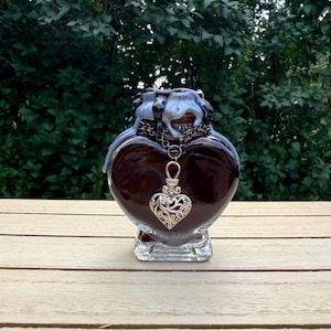 Peut inclure: Une bouteille en verre en forme de cœur avec un intérieur brun foncé et une base en verre transparent. Le haut de la bouteille présente un effet de goutte de cire bleu clair. Une chaîne en métal argenté entoure le goulot avec un pendentif cœur.