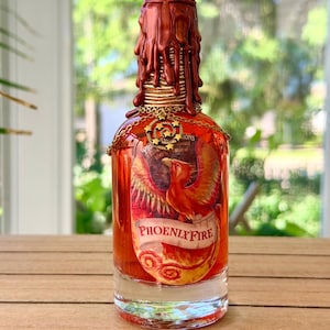 May include: A clear glass bottle filled with orange liquid, featuring a label with a phoenix illustration and the text PHOENIX FIRE. The neck is sealed with dripping metallic copper wax, and a gold-toned chain with a star charm hangs around it.