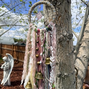 Peut inclure: Une suspension murale circulaire composée d'un cerceau enveloppé orné d'un cristal rose central, de rubans pendants dans des tons rose, blanc et vert, de clochettes en laiton, de dentelle blanche et de petites perles, suspendue par un ruban rose pâle.