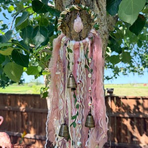 Peut inclure: Une suspension murale circulaire composée d'un cerceau enveloppé orné d'un cristal de quartz rose brut central. Des bandes de tissu rose et crème, des accents de vigne verte, des perles florales et quatre clochettes en laiton sont suspendus.