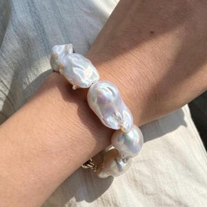 May include: A bracelet composed of several large, irregular white baroque pearls with a high-luster, iridescent finish. The pearls are strung together in a continuous row, showcasing their unique, organic shapes and smooth, reflective surfaces.