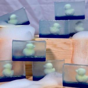 Pode incluir: Várias barras de sabão com base azul e topo transparente, cada uma contendo um pequeno patinho de borracha amarelo. Os sabonetes são dispostos sobre uma superfície de madeira com espuma branca, criando uma estética de spa.