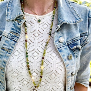 May include: A long beaded necklace featuring green, brown, and cream-coloured beads with small skull accents, worn over a white lace top and a light blue denim jacket. The text "Bones in the Forest" is visible at the top of the image.