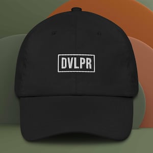 May include: Black baseball cap with a curved brim. The cap features the word "DVLPR" embroidered in white within a white rectangular border on the front. The cap is made of a soft fabric and has a classic, casual style.