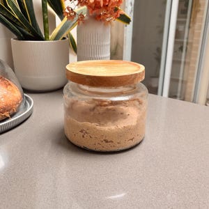 Puede incluir: Un tarro de vidrio transparente con tapa de madera, lleno de una sustancia marrón claro. El tarro está sobre una encimera gris. En el fondo, un jarrón blanco contiene flores naranjas y una planta en maceta es visible.