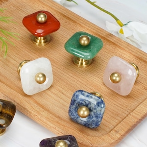 May include: A collection of seven square cabinet knobs made from various polished gemstones, including red, green, white, pink, brown, blue, and purple stones. Each knob features a central brass-toned metal stud and a matching brass-toned base.