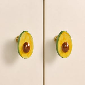 May include: Two cabinet knobs shaped like halved avocados. Each knob features a glossy green outer skin, a yellow-green flesh, and a central brown pit. The avocado-shaped fronts are attached to circular gold-toned metal bases on cream doors.
