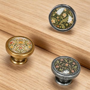 May include: Three round cabinet knobs with floral patterns under glass domes. One knob has a silver-toned metal base, one has a brass-toned metal base, and one has a dark, weathered grey metal base. Each features intricate botanical designs.