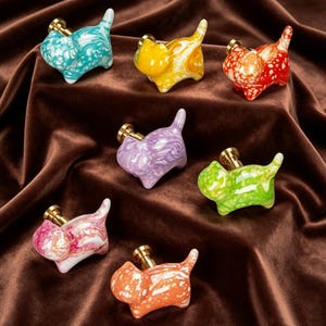 May include: A collection of seven cat-shaped cabinet knobs featuring a glossy, marbled finish in various colors including turquoise, yellow, red, purple, green, pink, and orange, each attached to a small, cylindrical brass-toned metal base.