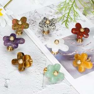 May include: A collection of flower-shaped cabinet knobs featuring various stone and resin materials in colors including green, purple, brown, white, red, and yellow. Each knob has a central brass-toned metal stud and a matching brass-toned base.