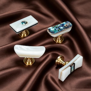 May include: Four cabinet knobs featuring brass bases and distinct designs. One is a white rectangle with a central iridescent inlay, one has a curved iridescent shell top, one is a smooth white curved piece, and one is a white bar with a central dark inlay.