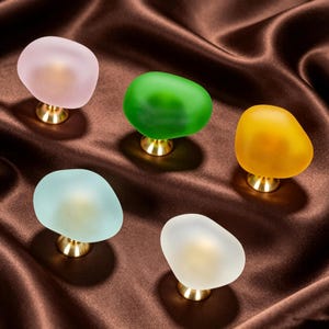 May include: Five cabinet knobs with irregular, pebble-shaped tops made of frosted glass in pink, green, yellow, light blue, and white. Each knob features a polished gold-tone metal base that connects the glass top to the mounting surface.