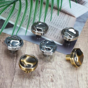 May include: A collection of five cabinet knobs with a rounded, irregular, and reflective surface. Three knobs feature a polished silver finish, while two knobs display a brushed, antique brass finish with dark, muted tones on the top surface.