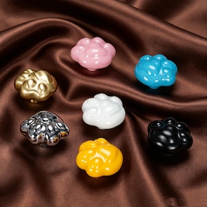 Peut inclure: Un ensemble de sept boutons de placard en forme de pattes d'animaux, avec des finitions variées incluant rose brillant, bleu vif, or métallique, chrome brillant, blanc uni, jaune éclatant et noir mat, chacun avec un design de patte arrondi.