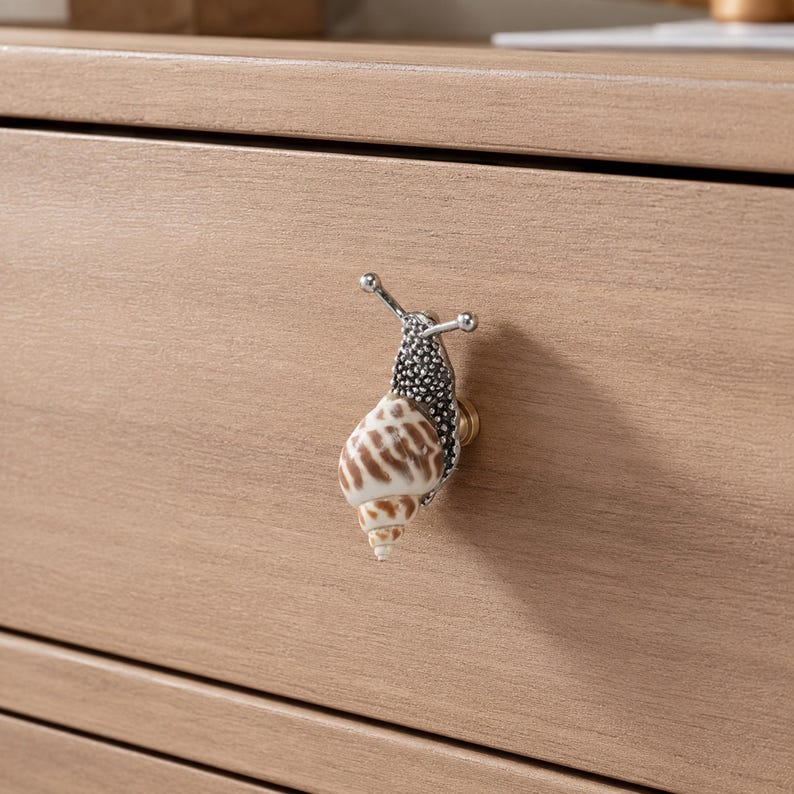 May include: A drawer knob shaped like a snail with a textured silver metal body and a natural spiral seashell for the shell. The snail features two silver antennae with small round tips and is mounted on a small gold-toned circular base.