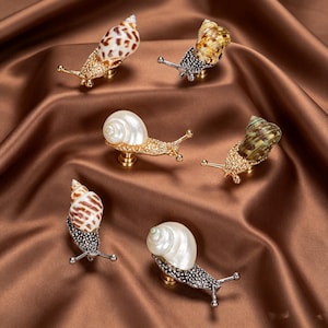 May include: A collection of six snail-shaped cabinet knobs featuring metallic bodies with textured surfaces and various natural seashell tops. The snail bodies are finished in gold or silver tones, each with two extended antennae and a mounting base.