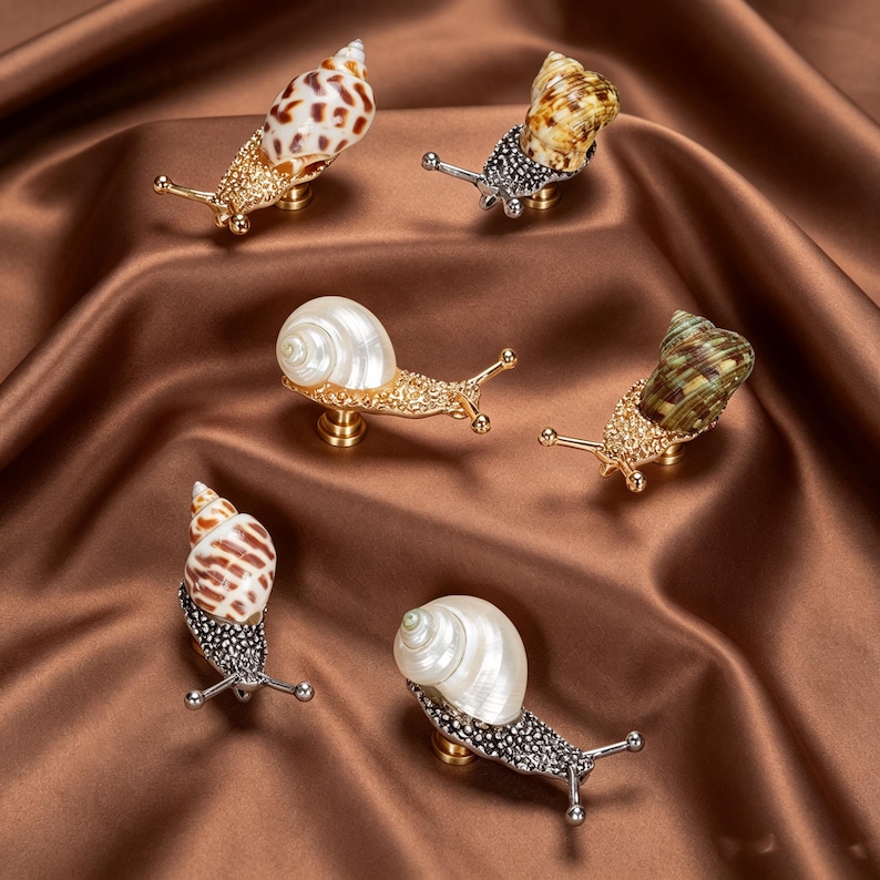 May include: A collection of six snail-shaped cabinet knobs featuring metallic bodies with textured surfaces and various natural seashell tops. The snail bodies are finished in gold or silver tones, each with two extended antennae and a mounting base.