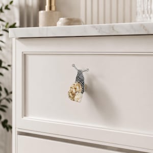 May include: A cabinet knob shaped like a snail with a textured metallic silver body and a natural spiral seashell shell. The snail features two protruding antennae and is mounted on a flat, off-white drawer front with a recessed panel design.