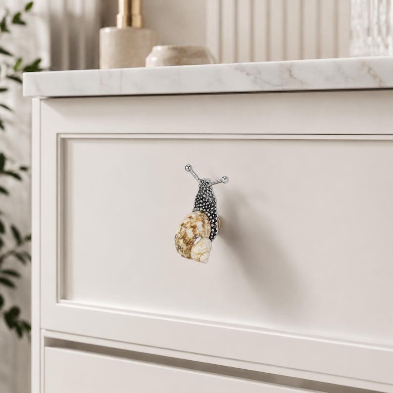 May include: A cabinet knob shaped like a snail with a textured metallic silver body and a natural spiral seashell shell. The snail features two protruding antennae and is mounted on a flat, off-white drawer front with a recessed panel design.