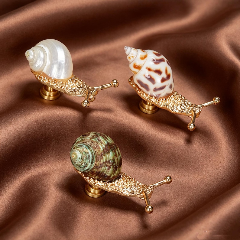 May include: Three snail-shaped cabinet knobs featuring gold-toned metal bodies with textured surfaces and distinct shells. One knob has a pearlescent white shell, one has a brown-spotted shell, and one has a green and brown striped shell.