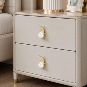 May include: A light beige two-drawer nightstand featuring two decorative drawer pulls shaped like snails. Each snail pull has a textured gold-toned metal body and a smooth, white, pearl-like shell. The nightstand has a smooth, matte finish.