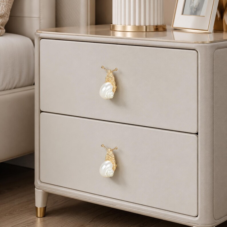 May include: A light beige two-drawer nightstand featuring two decorative drawer pulls shaped like snails. Each snail pull has a textured gold-toned metal body and a smooth, white, pearl-like shell. The nightstand has a smooth, matte finish.