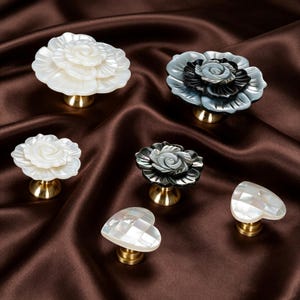 May include: A collection of six decorative cabinet knobs featuring floral and heart-shaped designs. The knobs are crafted from iridescent white and dark grey shell material, each mounted on a polished gold-tone metal base for furniture attachment.