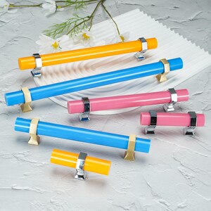 May include: A collection of cylindrical cabinet pulls in various lengths and colors, including bright yellow, vibrant blue, and soft pink. Each pull features metallic mounting brackets in either silver or gold tones, providing a secure attachment point for furniture installation.