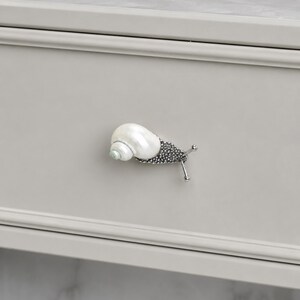 May include: A snail-shaped drawer knob featuring a white, iridescent seashell for the shell portion and a textured, dark metallic body with two thin, protruding antennae. The hardware is mounted on a light gray, flat-fronted drawer panel.