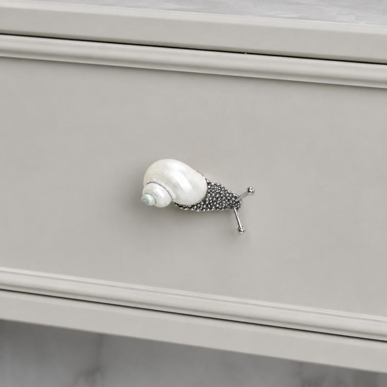 May include: A snail-shaped drawer knob featuring a white, iridescent seashell for the shell portion and a textured, dark metallic body with two thin, protruding antennae. The hardware is mounted on a light gray, flat-fronted drawer panel.