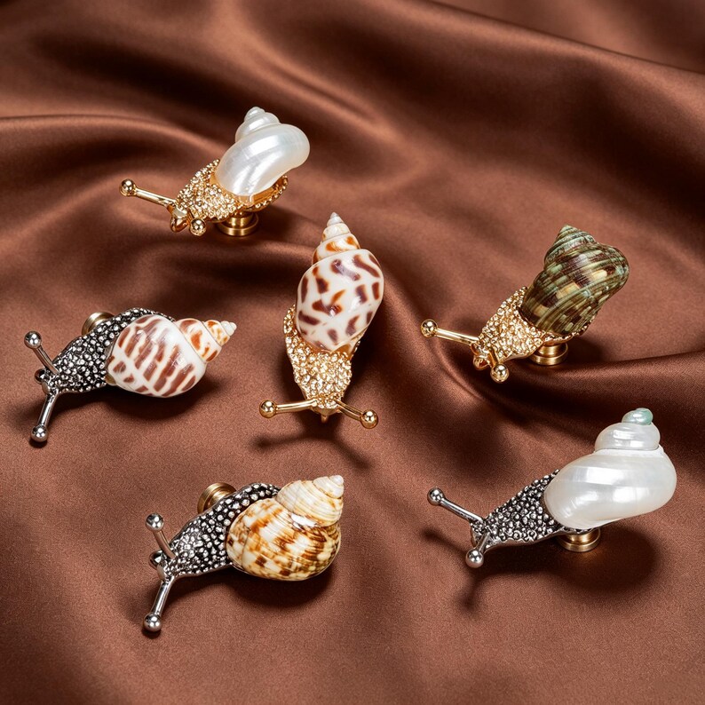 May include: A collection of five snail-shaped cabinet knobs. Each knob features a metallic body encrusted with small crystals, paired with a natural seashell acting as the snail shell. The designs include white, brown, and green shell variations.