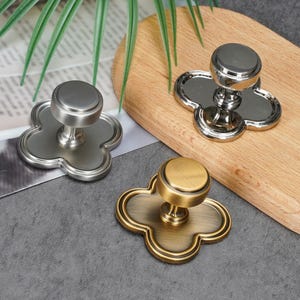 May include: Three metal cabinet knobs, each featuring a circular top and a clover-shaped base plate. One knob has a brushed silver finish, one has a polished chrome finish, and one has a brushed brass finish, all with a smooth, metallic texture.