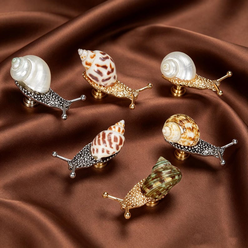 May include: A collection of six snail-shaped cabinet knobs featuring metallic bodies with textured surfaces and various natural seashell tops. The snail shells display diverse patterns and colors, including white, brown, and green, attached to gold or silver-toned metal bases.